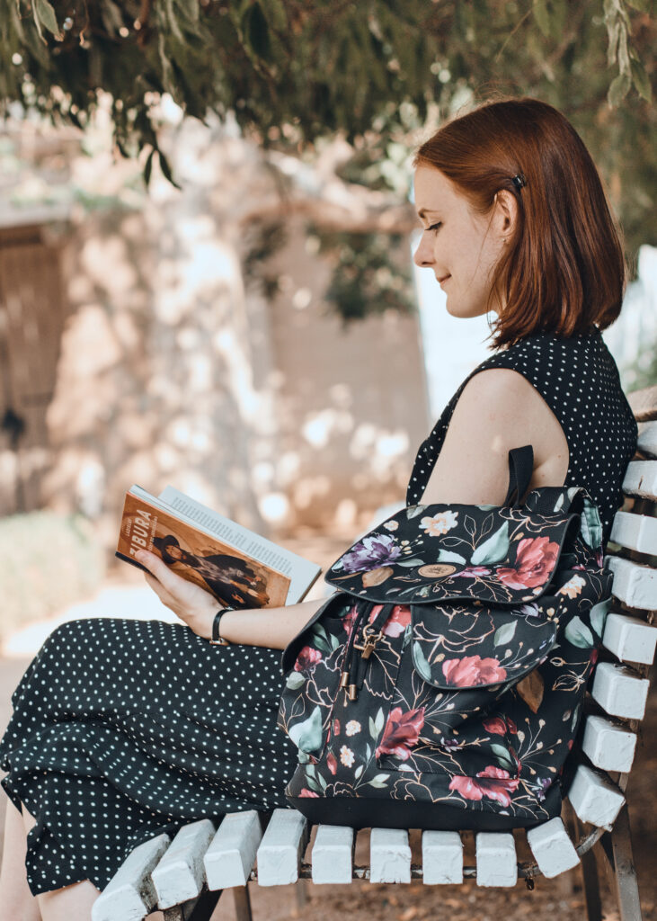 Žena sedící na lavičce čtoucí knihu s opřeným dámským květovaným batohem ELLIE Od Klárky
