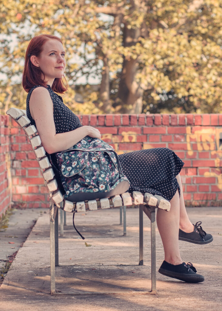 Žena sedící na lavičce v černých puntíkatých šatech s opřeným květovaným batohem ELLIE Od Klárky